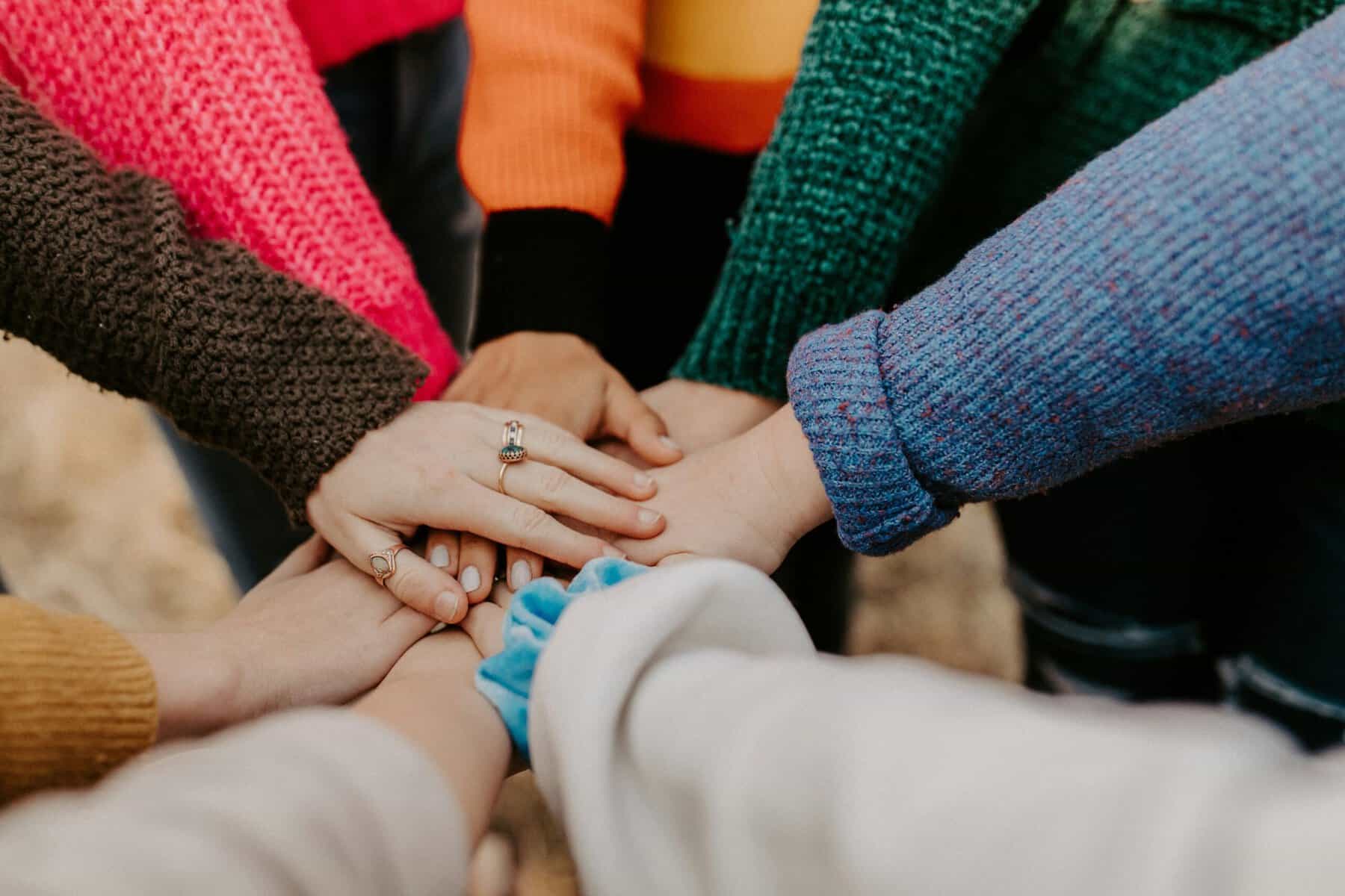 A group of women who have their hands connected over in the centre. There are 6 hands in the centre, representing unity and International Womens Day and support for women's mental health.