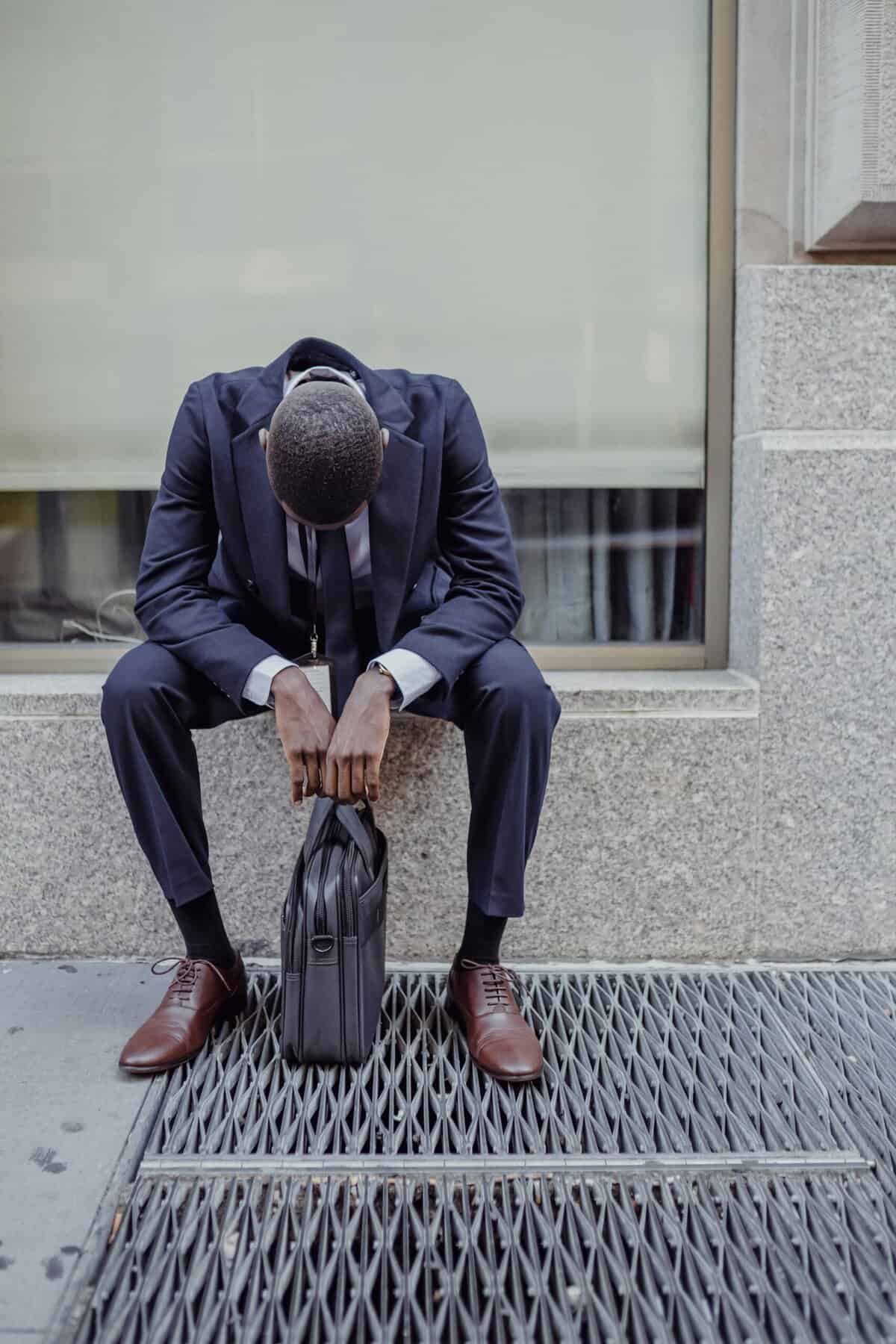 A man possibly experiencing burnout. He is sitting outside in a suit with his shoulders and head slumped into himself as a briefcase rests between his feet.