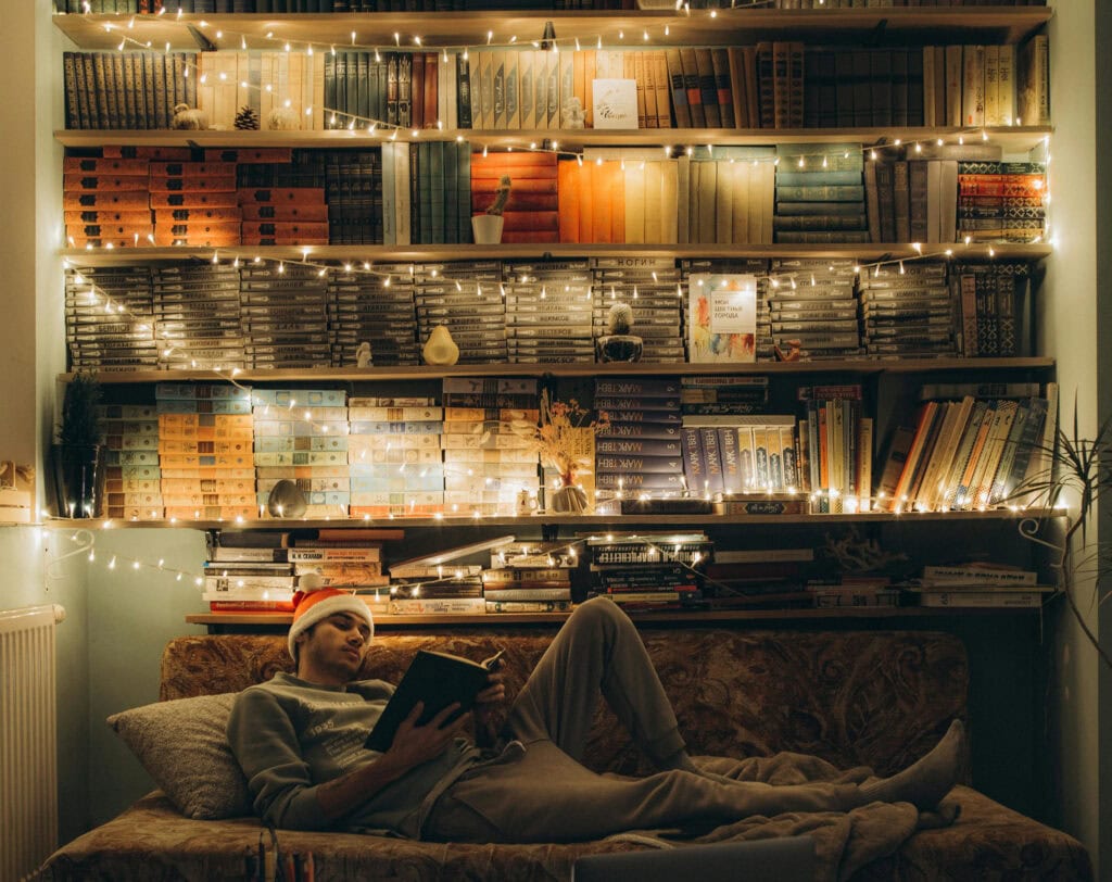 Man reclining on sofa lit with fairy lights on full bookshelves behind him, he wears a santa hat and is relaxing alone reading a book. It looks warm and cozy showing that even if you are alone this christmas, that can also be okay.