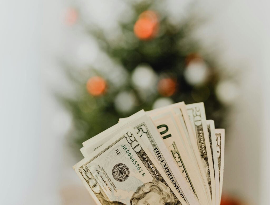 Foreground is a handful of dollar bills in front of a small christmas tree that is out of focus - representing the financial cost and sometimes associated stress of the holidays.