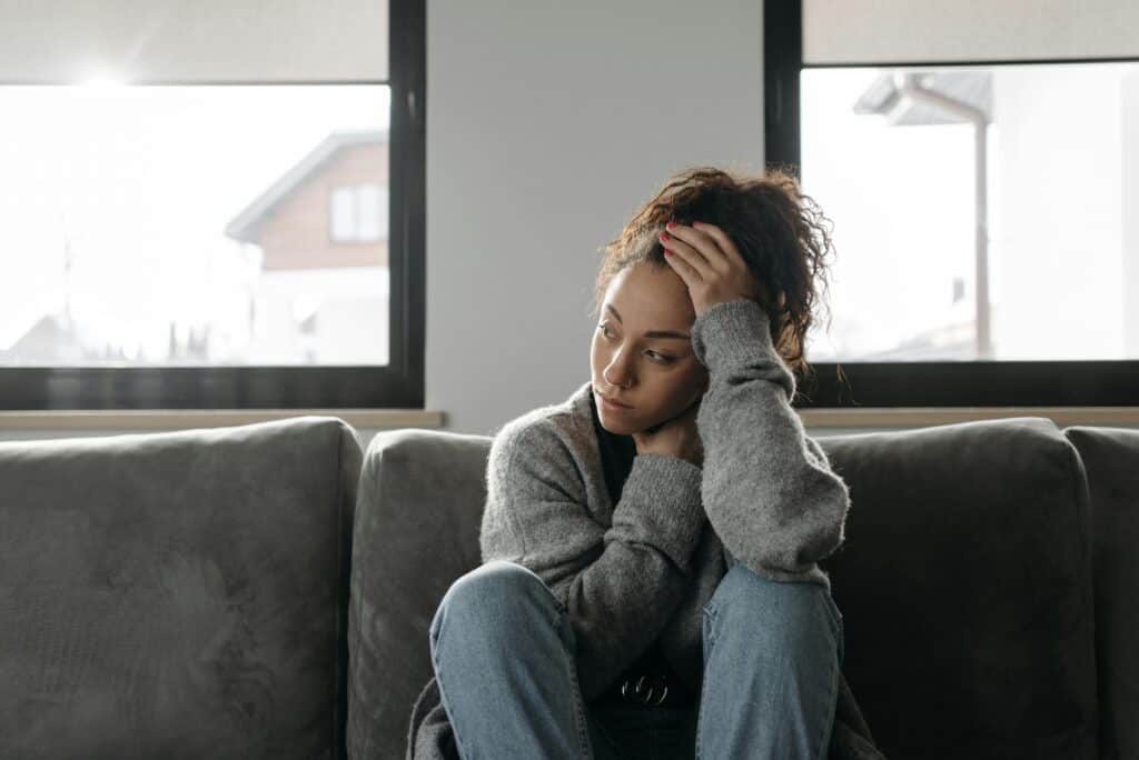 Young mixed race woman sitting alone on grey sofa with one hand on her forehead and one hand tucked in across her body. She looks despondent. This represents stressful family situation during the holidays, whether through conflict, grief, challenging behaviour, etc.
