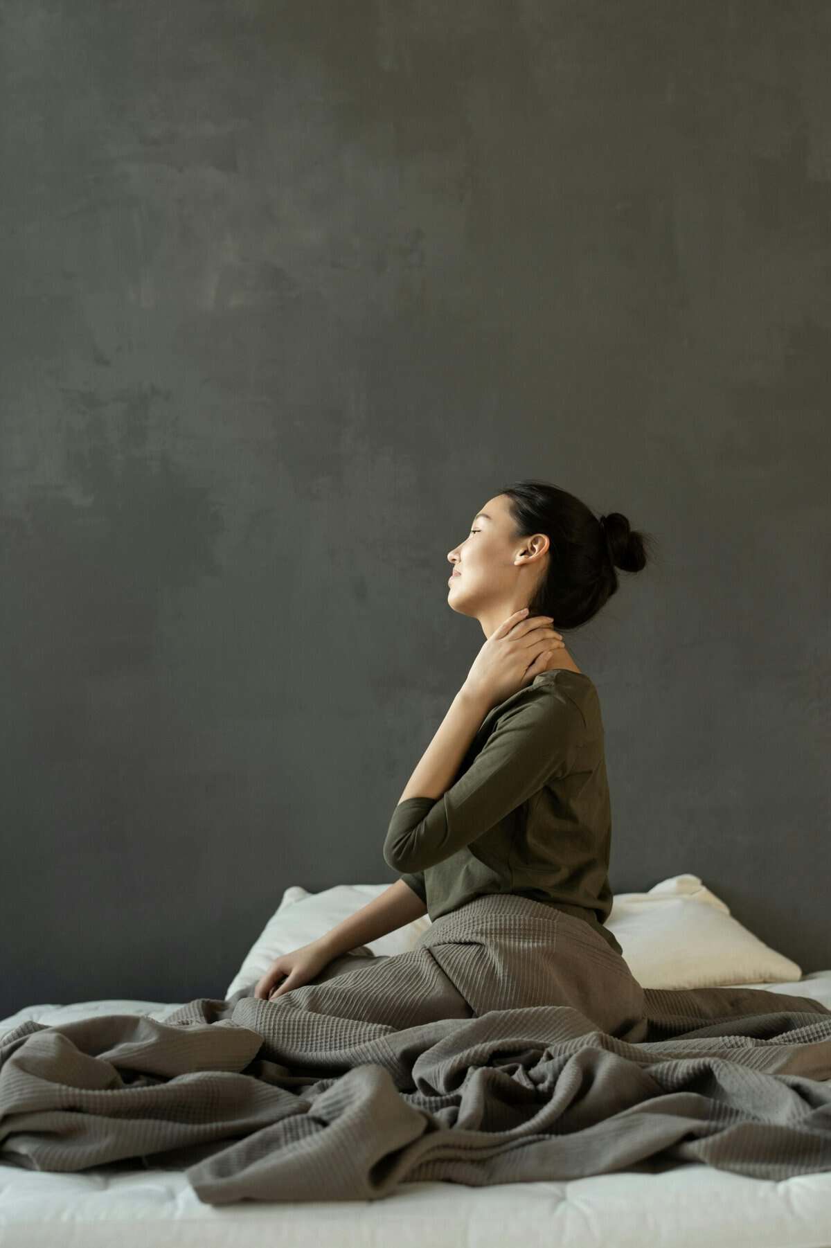 A woman sits quietly on a grey bed with her eyes closed, one hand resting on her neck. The image captures a moment of reflection, self-care, and the ongoing reality of living with chronic pain or physical discomfort.